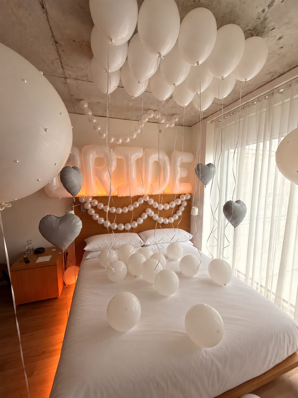 Hotel room styled for a bride with white ceiling balloons, pearl garland, silver hearts and a glowing BRIDE balloon banner