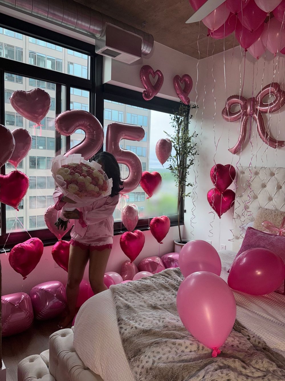 Birthday girl holding a rose bouquet in a room filled with pink heart balloons, 25 number balloons and a pink bow balloon
